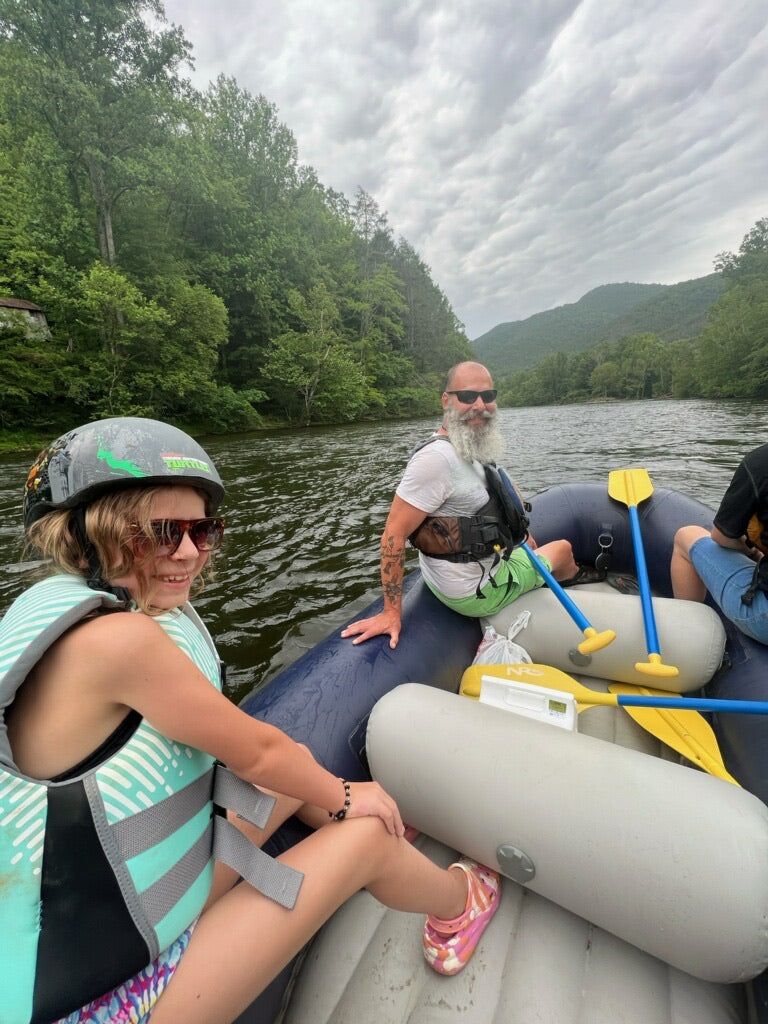 a middle aged man with a white beard sitting in a white water raft next to a young girl wearing a blue life jacket and helmet. they are both floating down a river in front of tall green mountains