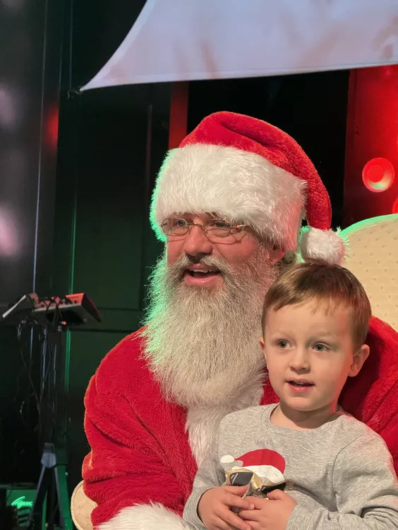 a middle aged man dressed as santa with a large white beard sitting on a cream colored plush chair and a young boy sitting on his lap. they are both smiling and sitting in front of a stage with dark walls and red lights