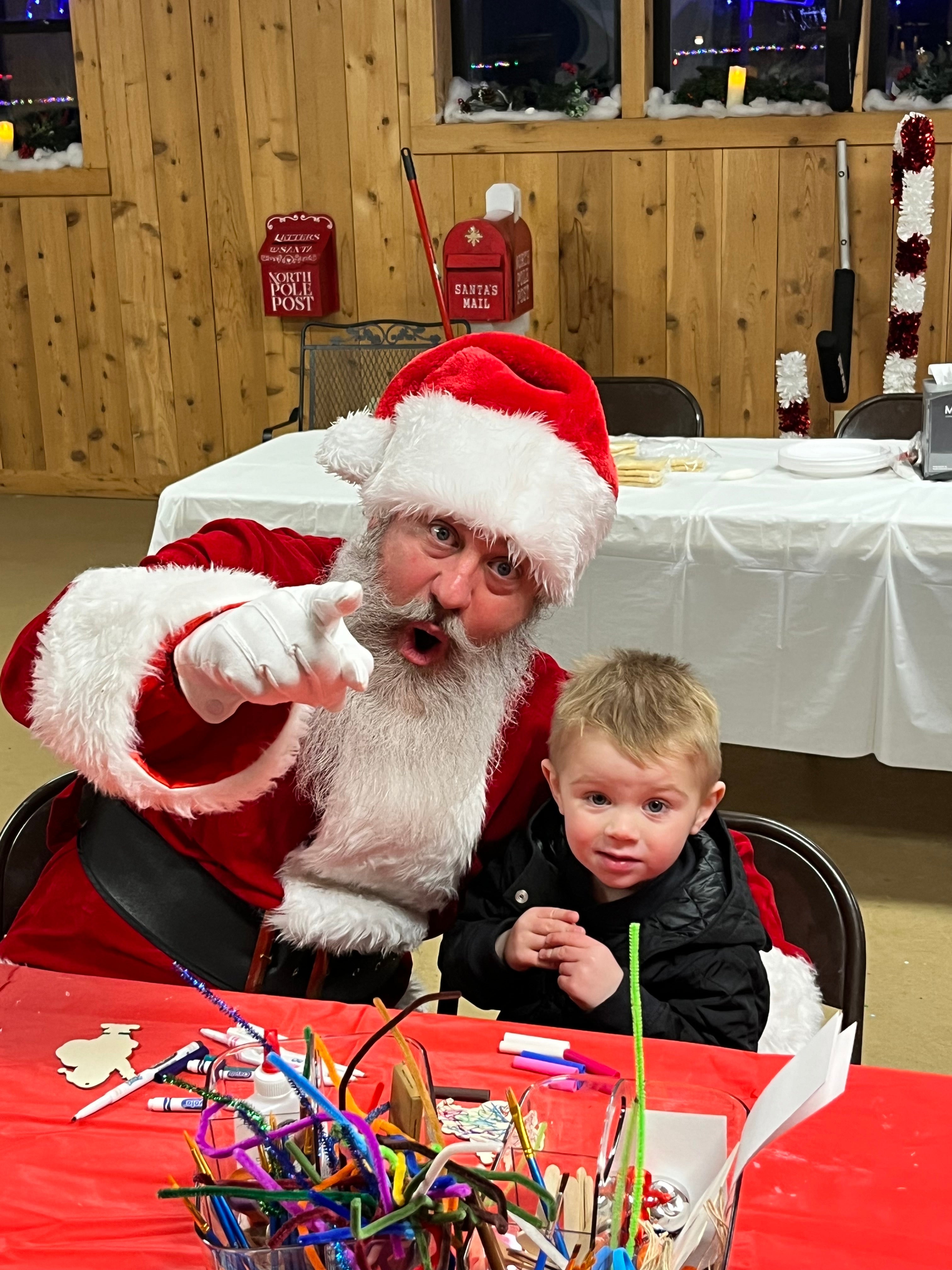 a middle aged man with a large white beard dressed as santa claus has his mouth wide open and points at the camera making a face. he is sitting next to a young boy making a craft with him. they are seated at a table with a red table cloth, and in front of a table with a white table cloth and in a room with brown wooden walls and tan tile floors