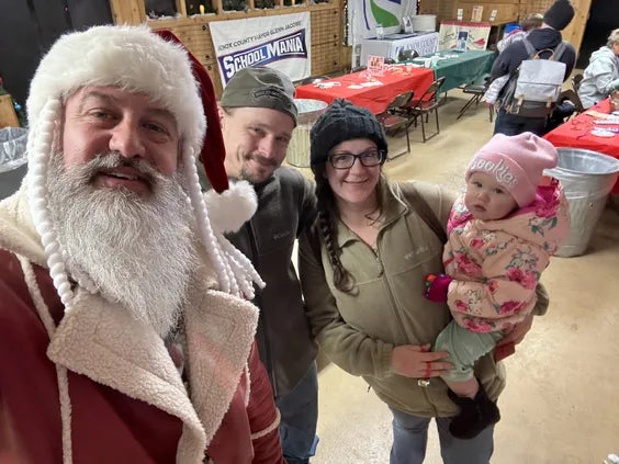 a middle aged man dressed as santa with a red leather coat and a large white beard standing next to two adults and one baby smiling in  a room with wooden walls and tables set up wiht red and green tablecloths