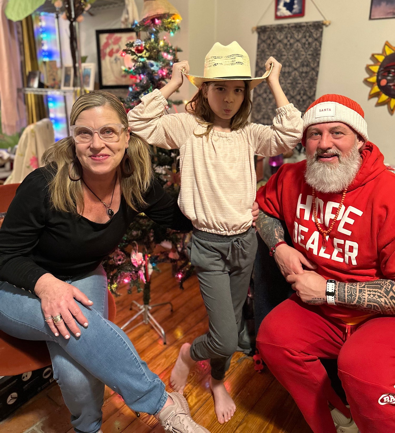 an image of three people sitting in a living room at christmas time. the mother is on the left wearing blue jeans, a black shirt, black earrings and necklace, pigtails in her hair, and clelar glasses. the child in the middle is wearing gray pants, a light colored shirt, and a cowboy hat. the person on the right is wearing all red with a hoodie that reads "hope dealer" and a red hat with the word "santa" on the front. he has tattoos up his arms and a fully white beard and an orange neckalce.