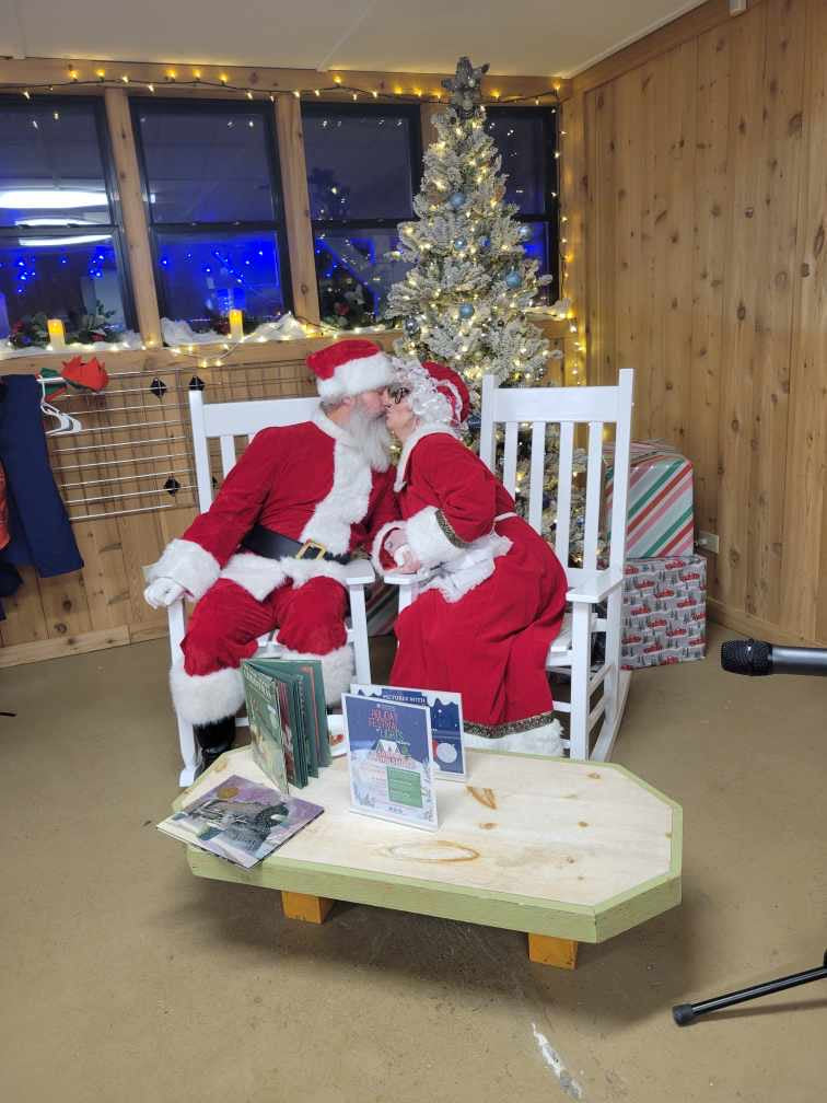 a middle aged man dressed as santa claus with a large white beard sitting in a rocking chair kisses a middle aged woman dressed as mrs. claus also sitting in a white rocking chair. the room behind them is decorated in a christmas theme, and includes a large christmas tree behind the people