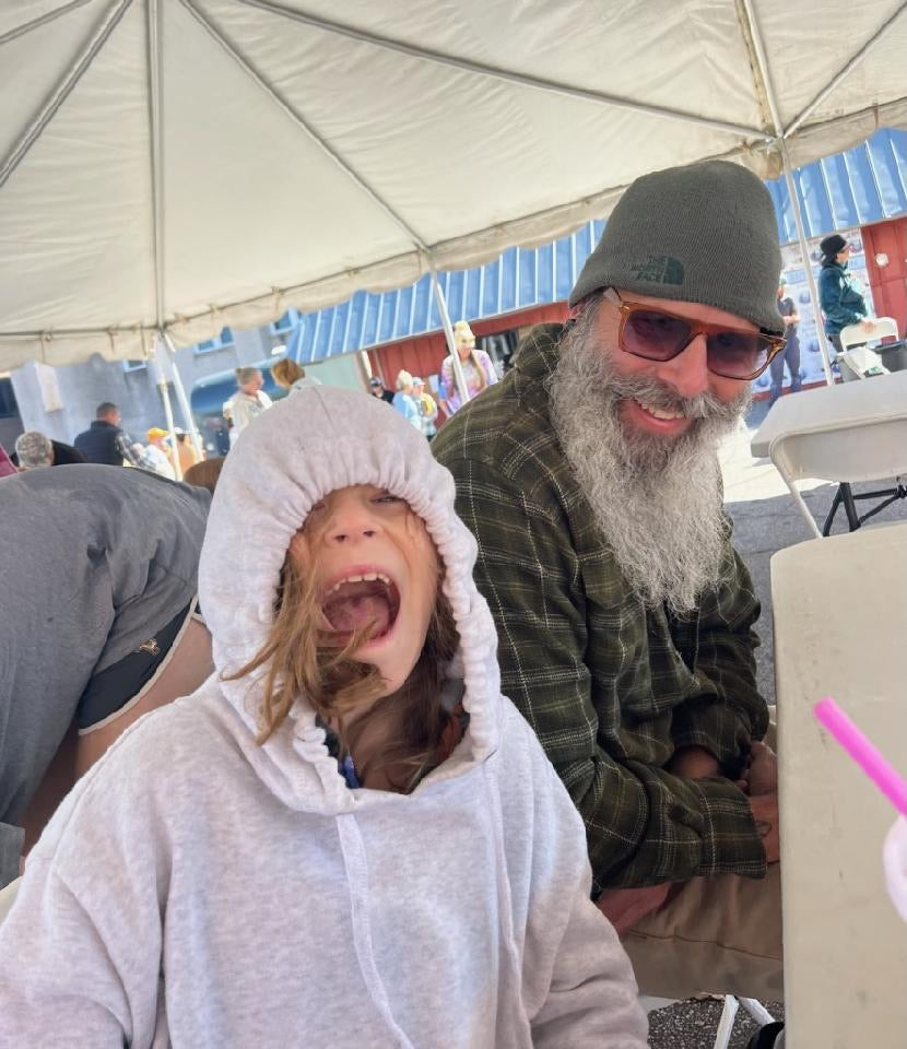 a middle aged man with a white beard smiling at a young girl smiling next to him with a gray hoodie sitting under a tent in front of a building with a gray roof