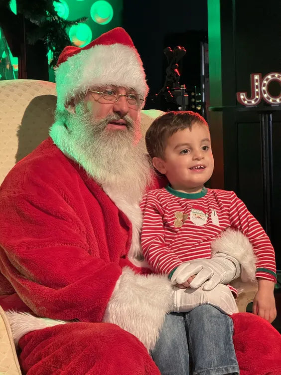 a middle aged man dressed as santa with a large white beard sitting on a cream colored plush chair with a young boy on his lap. the boy is wearing a christmas themed outfit and thehy are both sitting in front of a stage with dark walls and green lights. 