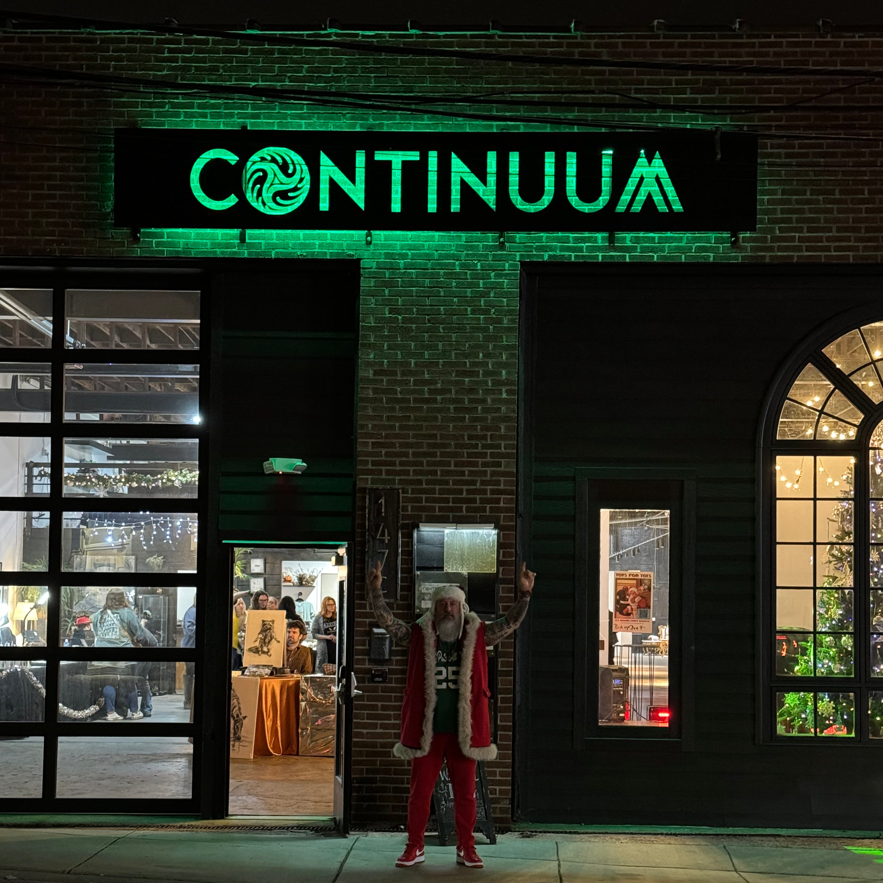 A middle aged man dressed in a christmas themed outfit and a large white beard and in front of a brick building with a large green neon sign that says "continuum." he is pointing up at the sign.