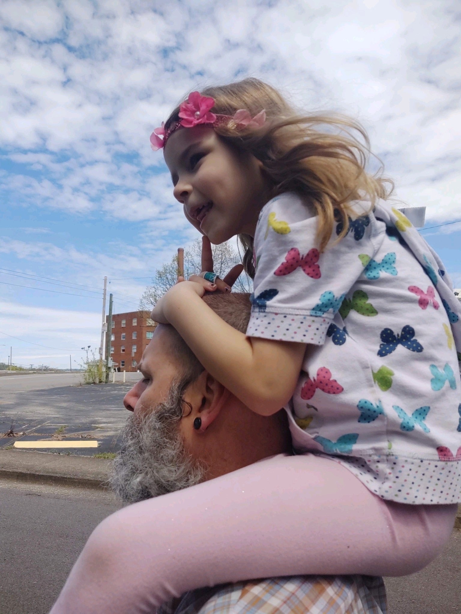 A middle aged man with a white beard and a young girl with flowers in her hair on the mans shoulders in front of a blue sky and an industrial looking building