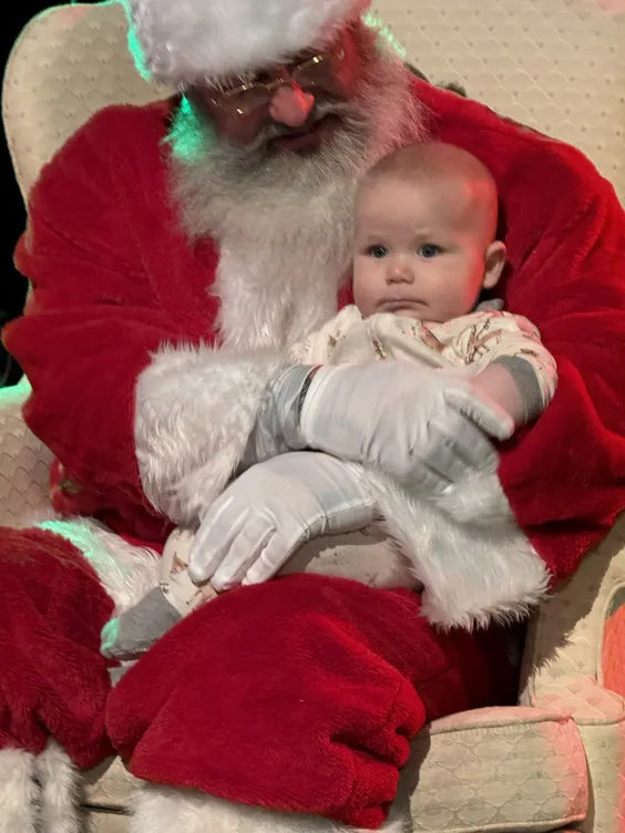 a middle aged man dressed as santa with a large white beard sitting on a cream colored plush chair with his arms wrapped around a young baby sat on his lap. 