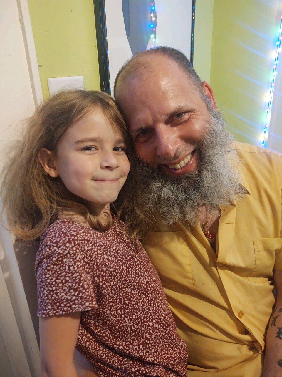 A middle aged man with a white beard holding a young girl in his arms with LED lights behind them. the girl is wearing a red and white spotted shirt and the man is wearing a yellow shirt.