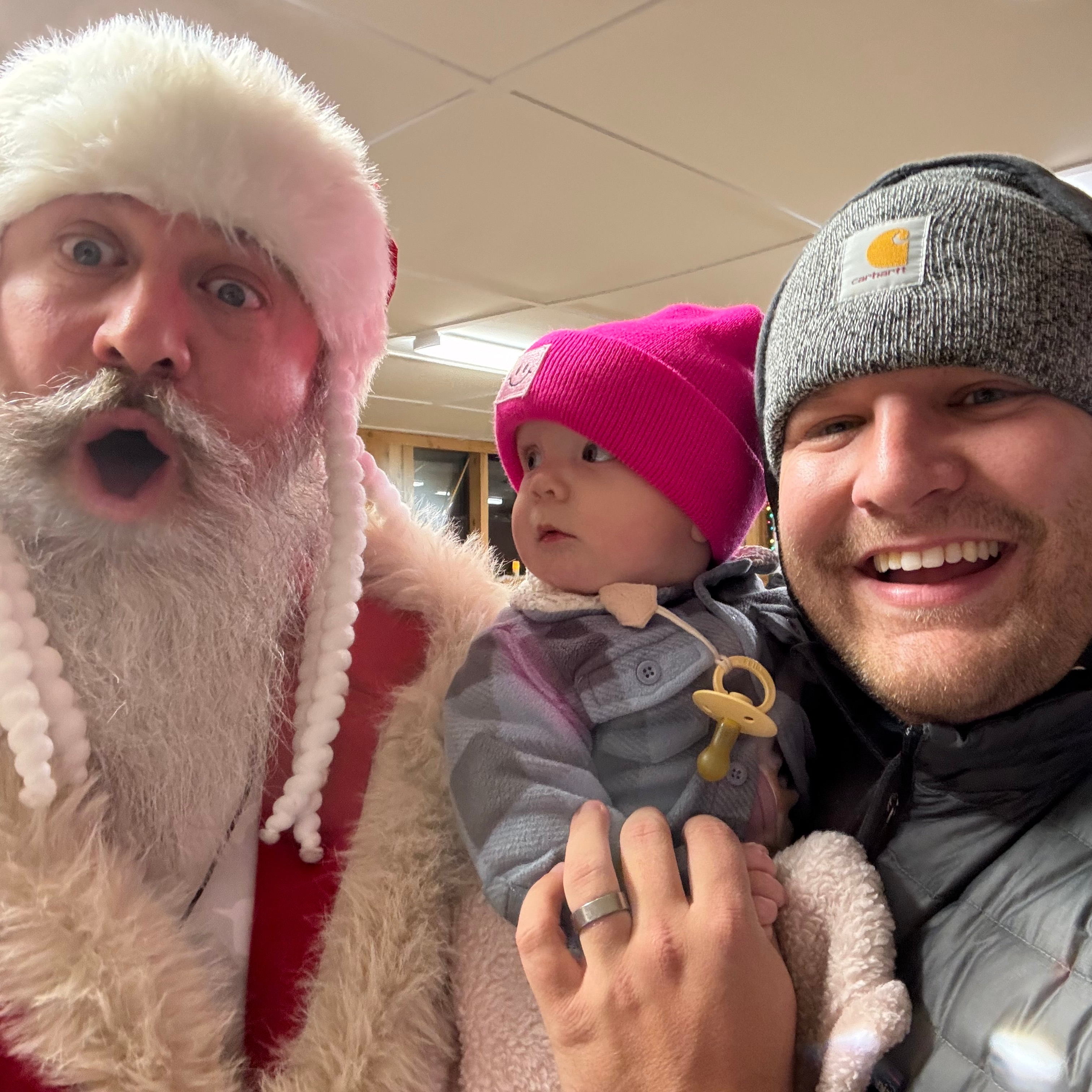 a middle aged man dressed as santa with a large white beard and his mouth wide open makes a face at the camera. the man next to him is smiling in warm clothes, and the baby the man is holding is looking at the man dressed as santa