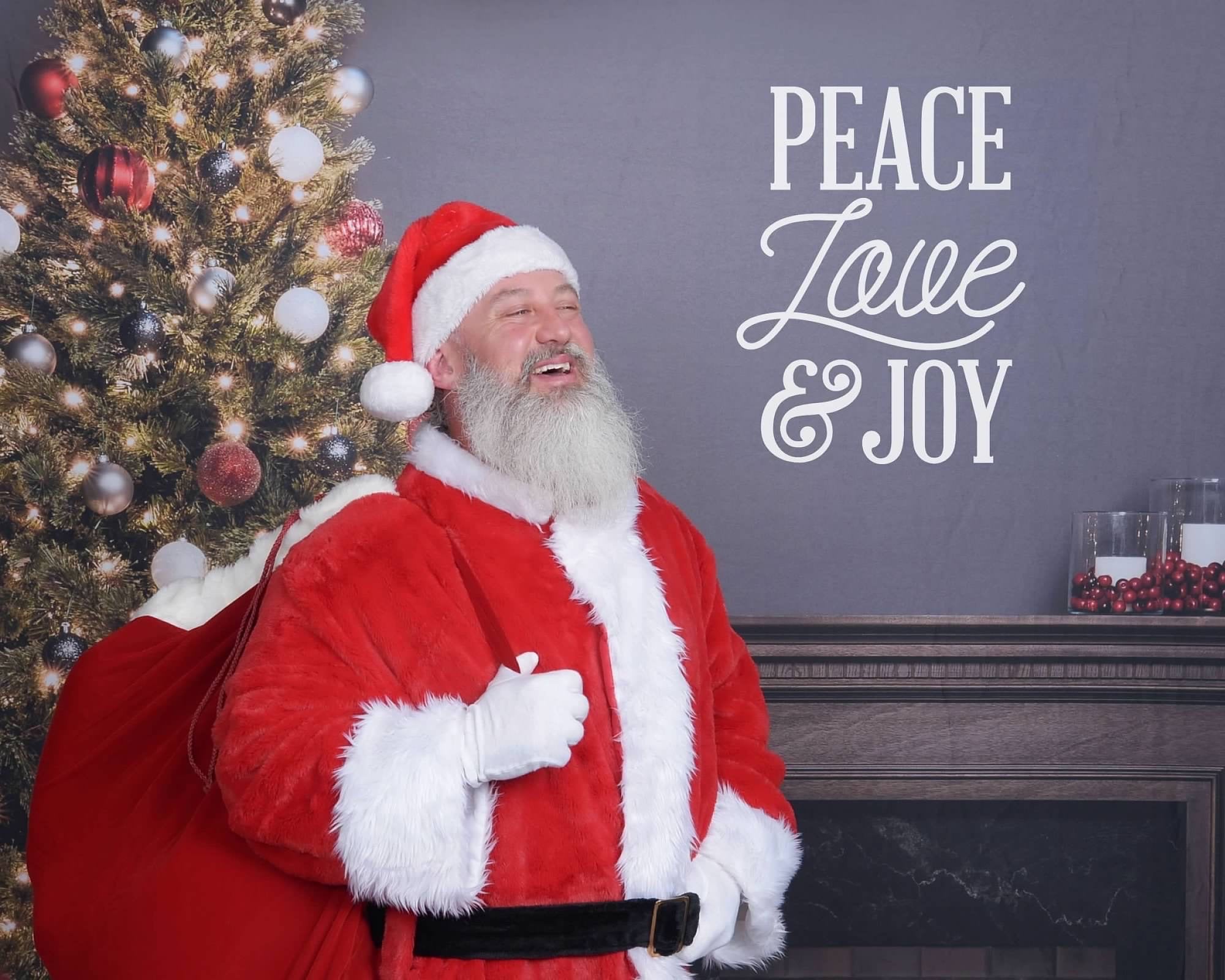 a middle aged man with a large white beard laughs in a jolly way, holding a sack of toys standing in front of a fireplace, christmas tree, and text on the wall that says "PEACE LOVE & JOY"