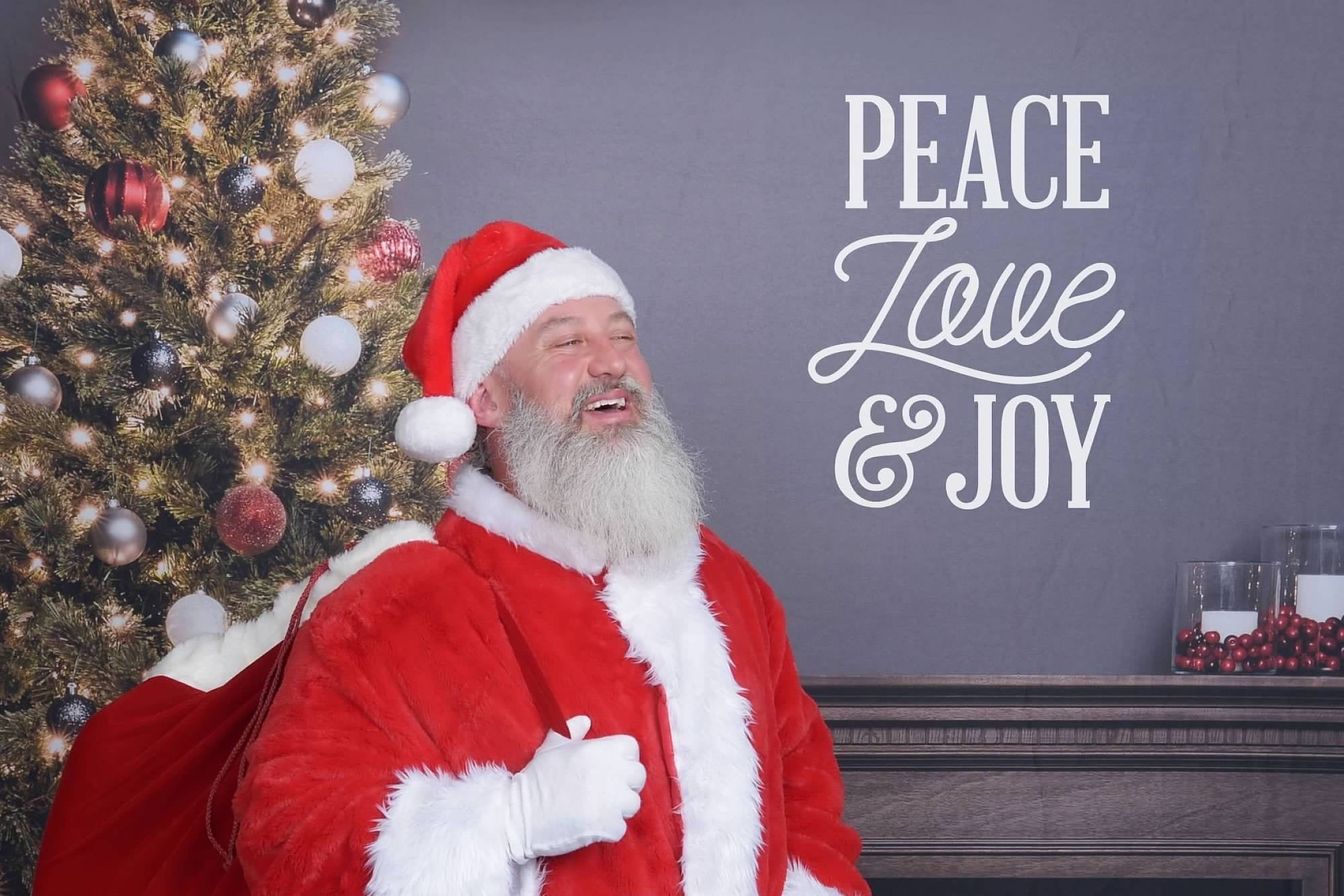 A Jolly looking man dressed as santa claus is holding a sack of toys in front of a  gray background with a fireplace, a christmas tree, and text on the wall that reads "peace love & joy"
