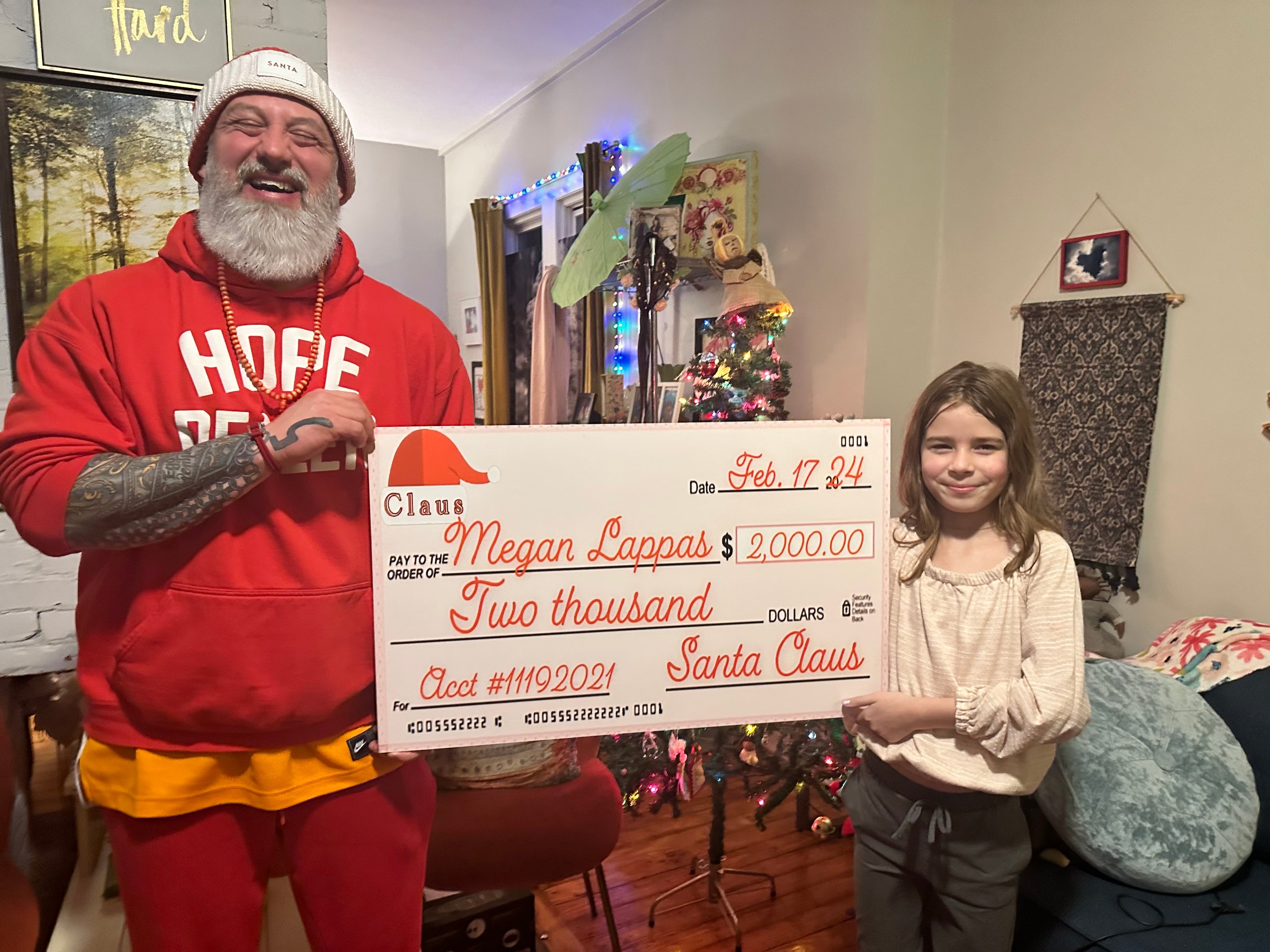 a middle aged man dressed in a christmas outfit holding a check with a young girl in a christmas themed living room