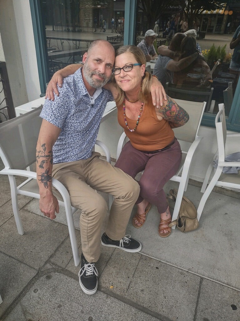 A middle aged man with a short beard with his arm around a middle aged woman sitting outside at a restaraunt