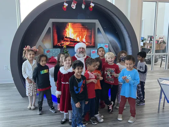 a middle aged man with a large white beard dressed as santa claus sitting in front of a space themed backdrop with an artificial fireplace. around him are lots of kids in christmas themed outfits and smiling