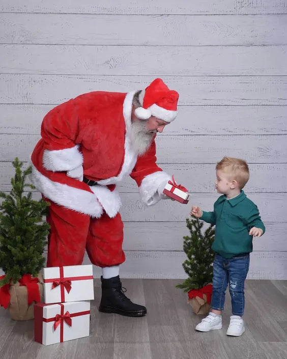 a middle aged man dressed as santa claus with a large white beard bending over handing a young boy a gift box. they are standing in front of a white wall with wooden panels and have small christmas trees around them. 