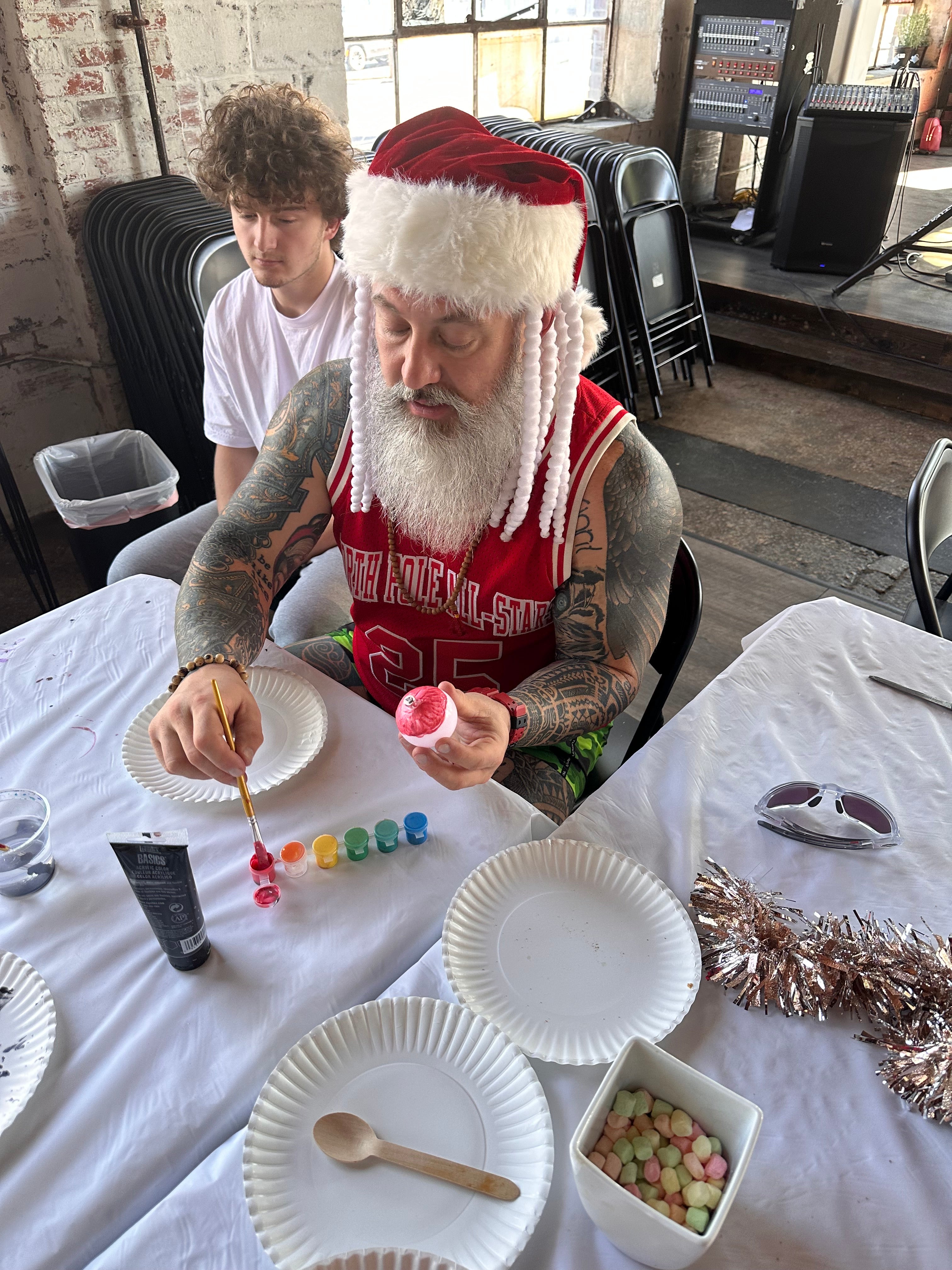 a middle aged muscular man dressed as santa claus with tattoos and a santa hat is painting a craft at a christmas themed event. the room in the background contains a stage, stone floors, and brick walls