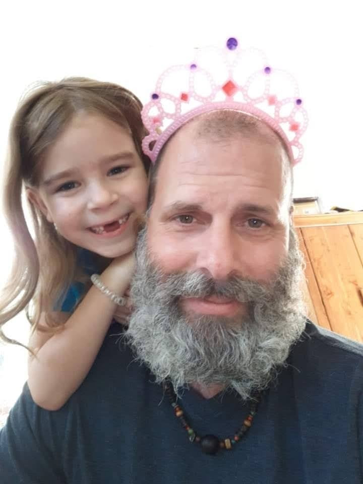 a middle aged man wearing a pink tiara and a white beard and a young girl peeking over his shoulder smiling in front of a wooden cabinet