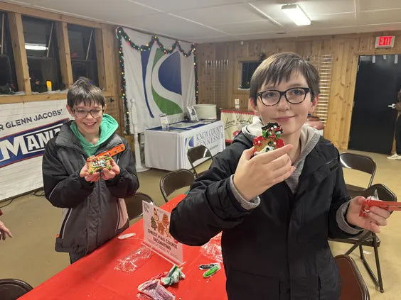 two boys holding christmas decorated cookies at a christmas themed event. they are in a room with a white roof and   walls with light wood grain. in the background there is a table with a red table cloth and a table with a white tablecloth with the "knox county parks and recreation" logo on it