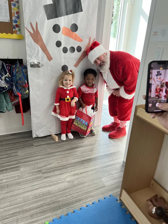 a middle aged man dressed as santa with a white beard crouching next to two young children also in christmas themed outfits. the kids are standing in a school in front of a door decorated like a snowman. 