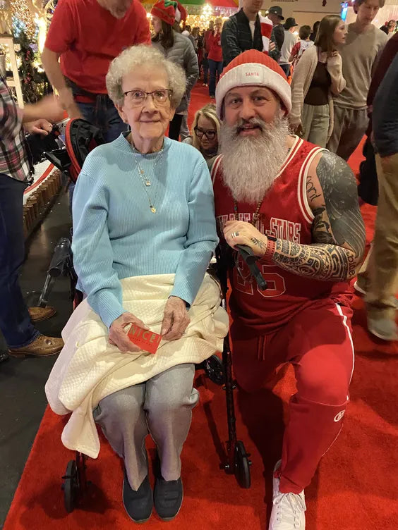 a middle aged muscular man dressed in a christmas themed outfit kneeilng next to an elderly woman. they are both smiling and at a christmas event