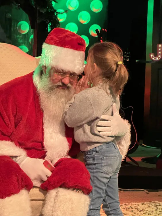 a middle aged man dressed as santa with a large white beard sitting on a cream colored plush chair with a young girl whispering something in his ear. the young girl is wearing jeans and a gray sweater, and they are both in front of a dark stage with a guitar behind them