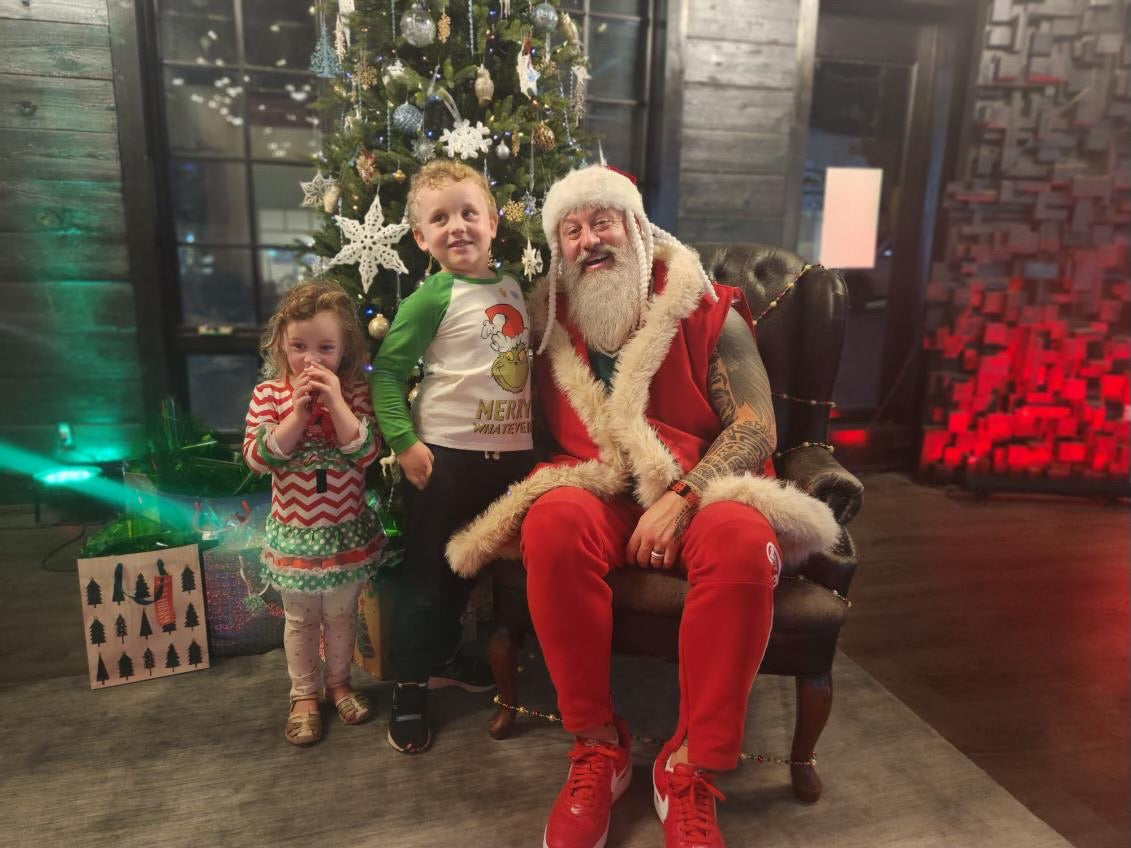 a middle aged man dressed in a christmas themed outfit sitting in a chair next to two standing young children. they are also dressed in christmas themed outfits. the room in the background is also christmas themed, with a large christmas tree directly behind the people