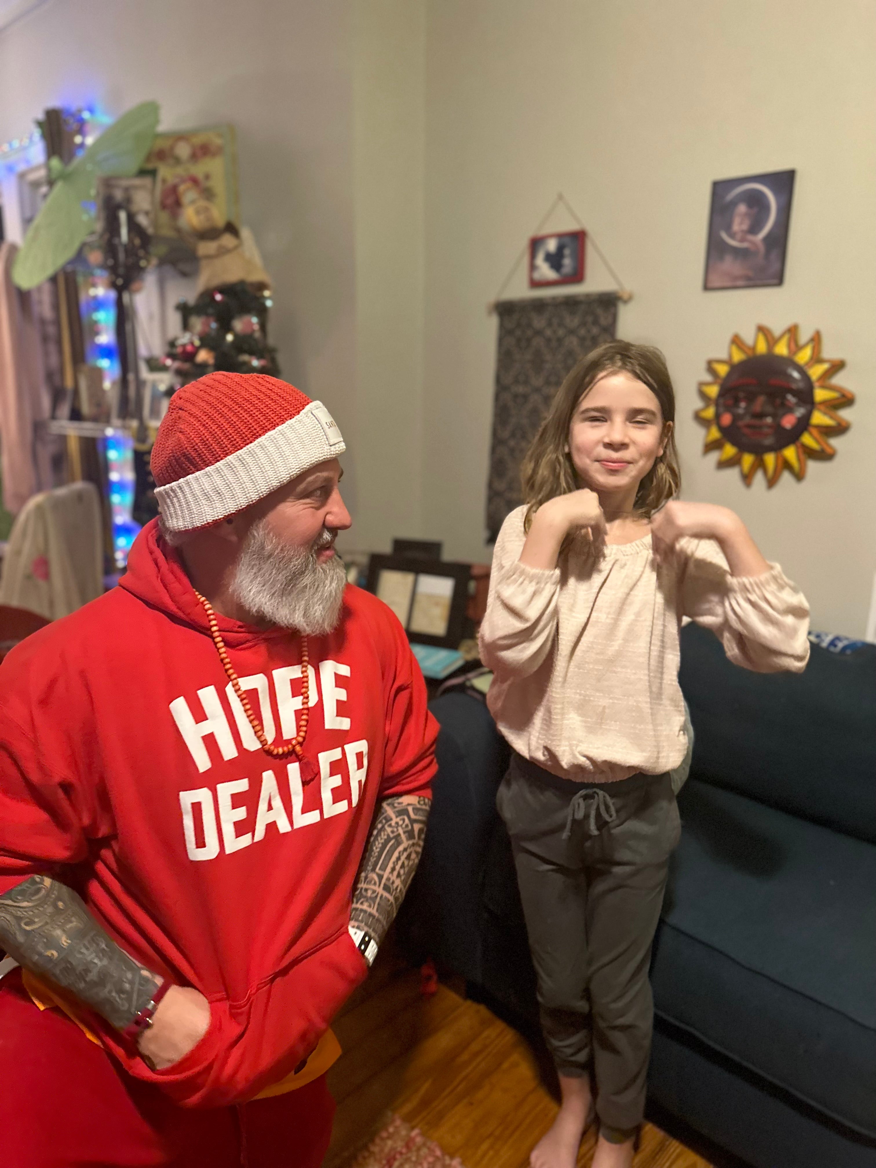 a middle aged man dressed in a christmas themed outfit kneeling next to a young girl in a living room.