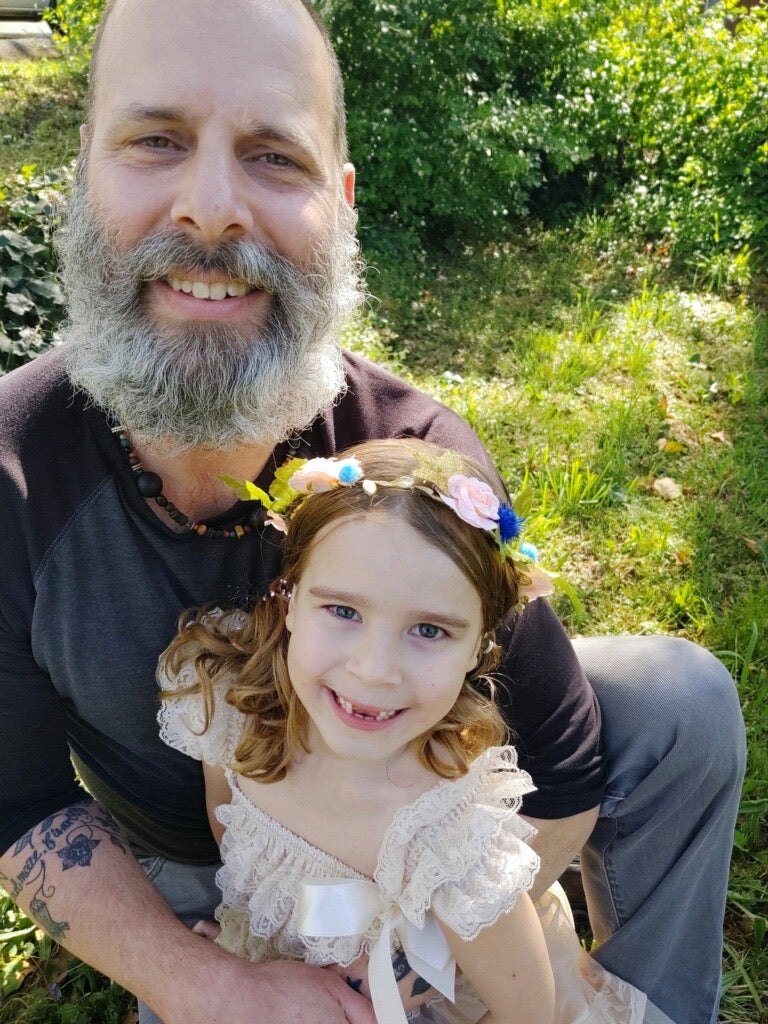 A middle aged man and a young girl standing in a patch of green shrubbery. the man has a full beard and a bald head, wearing gray pants and a black shirt. the girl is wearing a white dress, with flowers in her hair being embraced by her father.