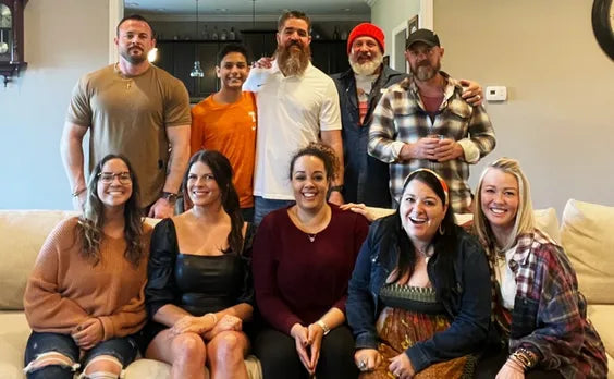 a group of people in an inviting beige living room. the women are sitting on a couch smiling, and the men, many of whom have beards, are standing behind the couch smiling.