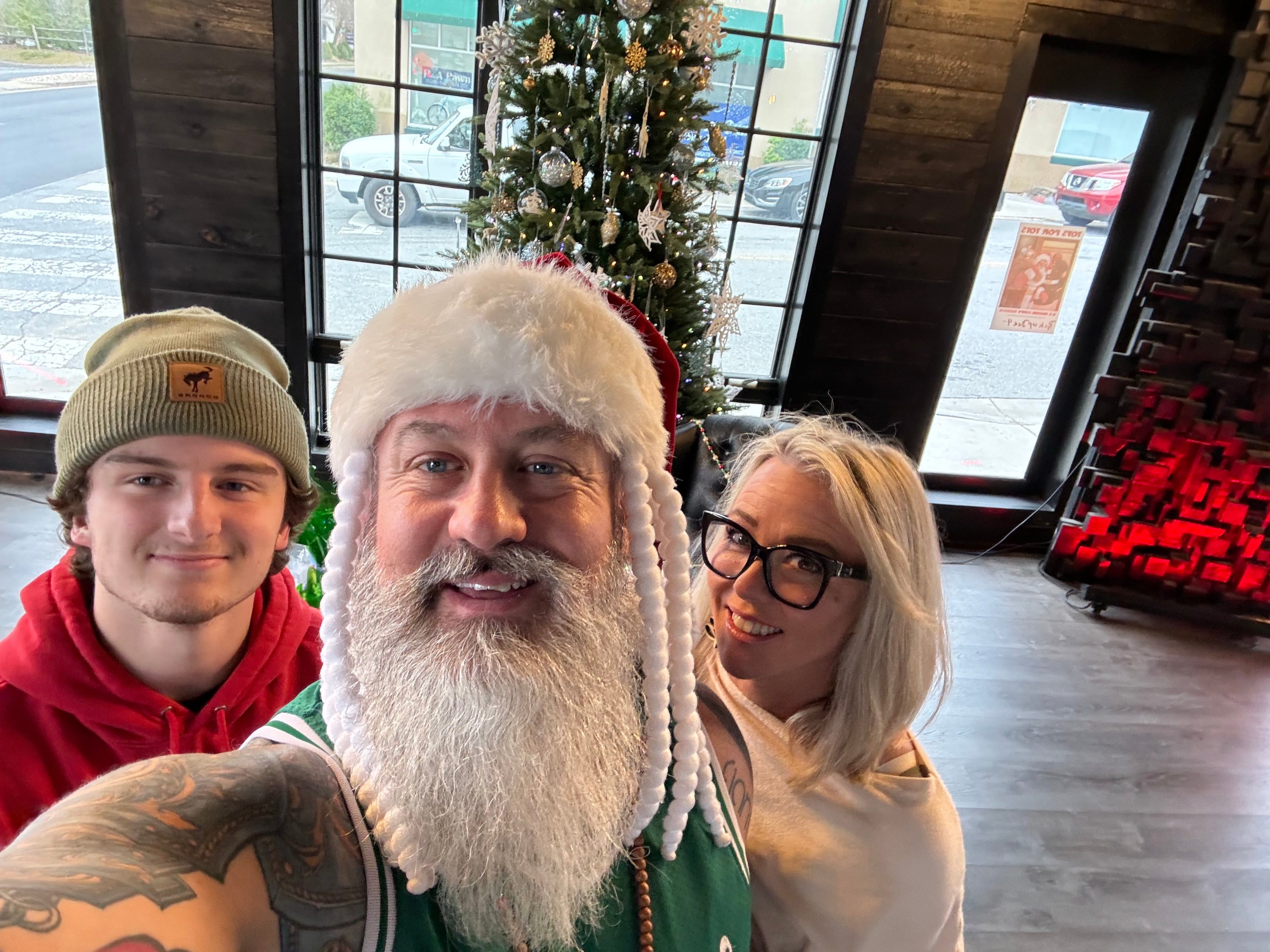 a middle aged man with a large white beard wearing a green chrsitmas themed jersey standing next to a middle aged woman and a younger man. they are all standing in a room with wooden walls and a christmas tree in the background. 