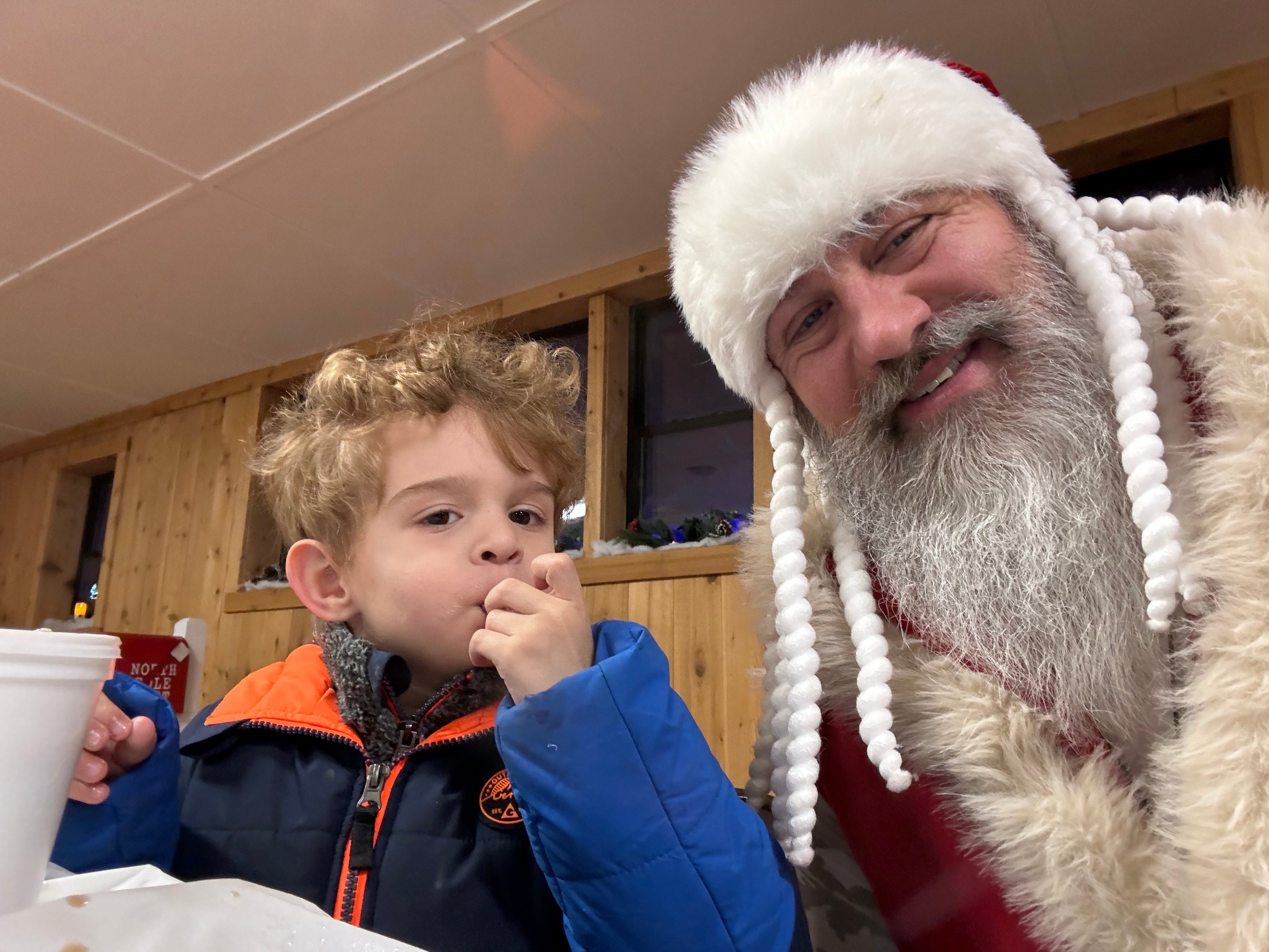 a middle aged man with a full white beard dressed as santa claus sitting next to a young boy eating something. they are both smiling and sitting in a room with brown wooden walls and a white roof. 