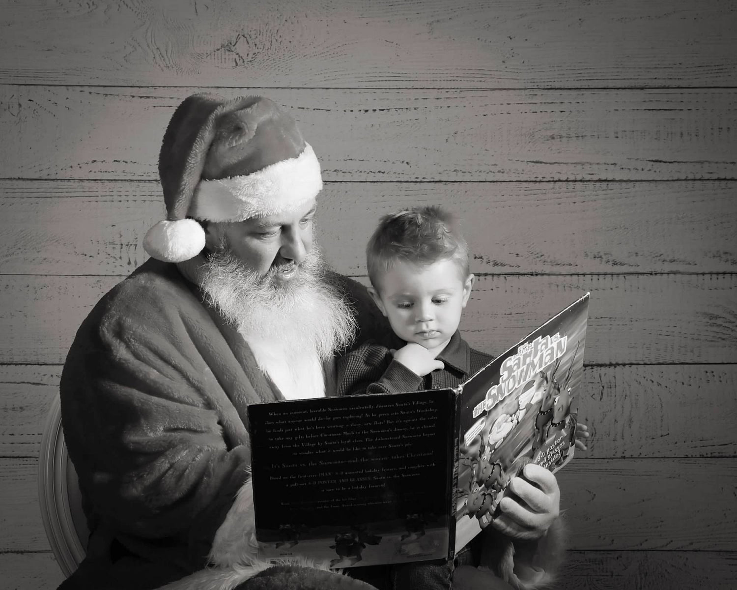 A black and white image of a man with a full white beard wearing a santa outfit with a small child sitting on his lap. Both of them are sitting in a chair and the background is made of wooden boards. he is reading a book to the child titled santa and the snowman. 