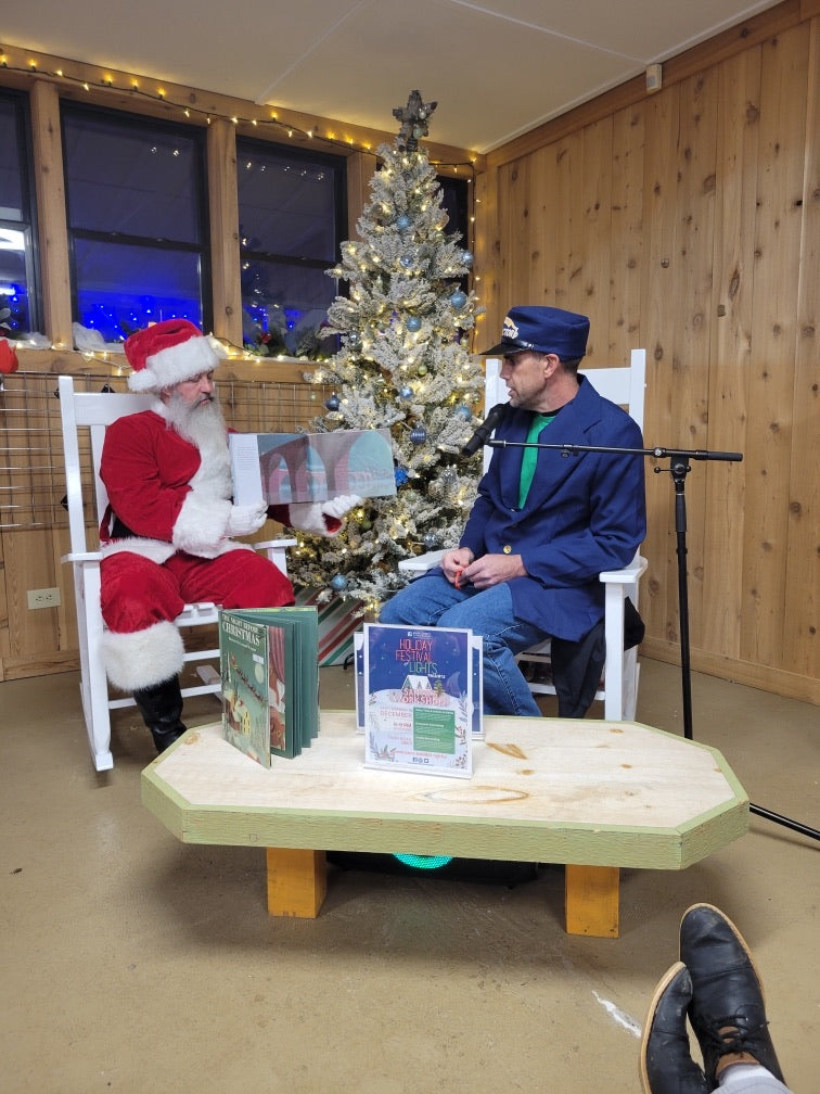 a middle aged man dressed as santa claus with a large white beard sitting in a white rocking chair preforming an interview with a man in a royal blue suit. the man is also sat in a white rocking chair and they are in a room with brown wooden walls, a white roof, and a large christmas tree behind them. 