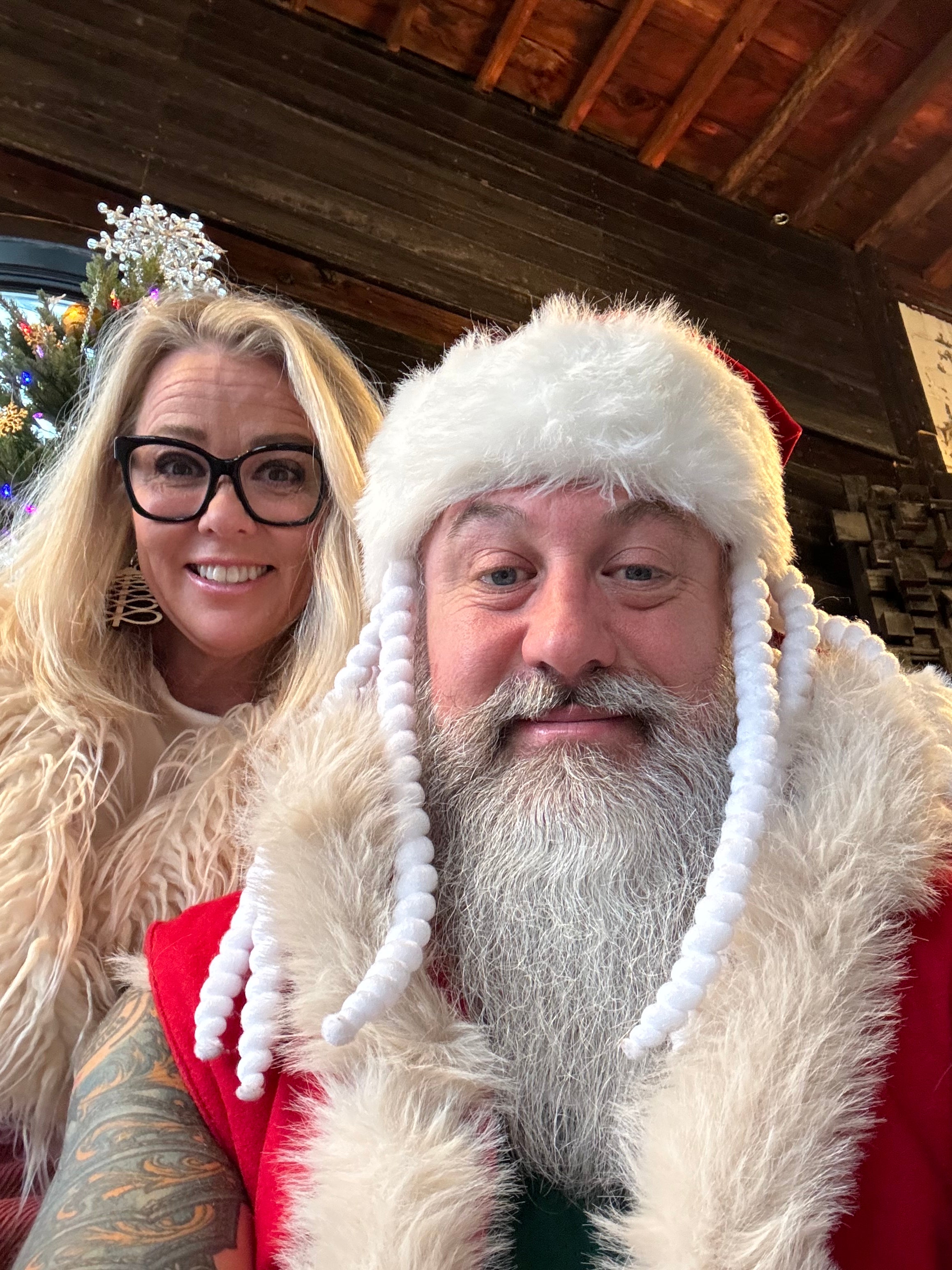 a middle aged man with a large white beard dressed as santa claus and a middle aged woman peering over his shoulder. they are both smiling and in a room that is themed like a log cabin