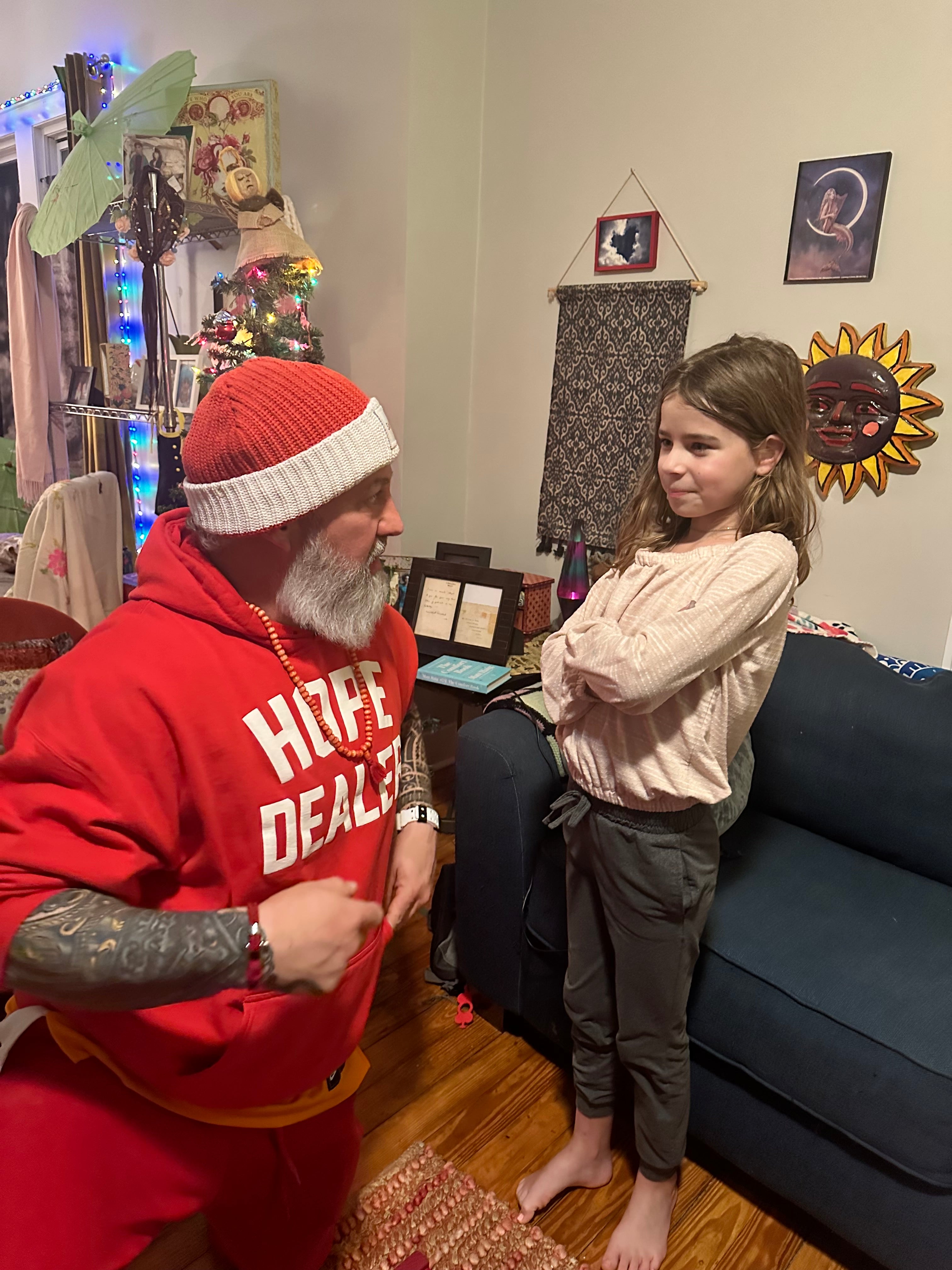 A middle aged man in a christmas themed outfit kneeling next to a young girl in a living room. 