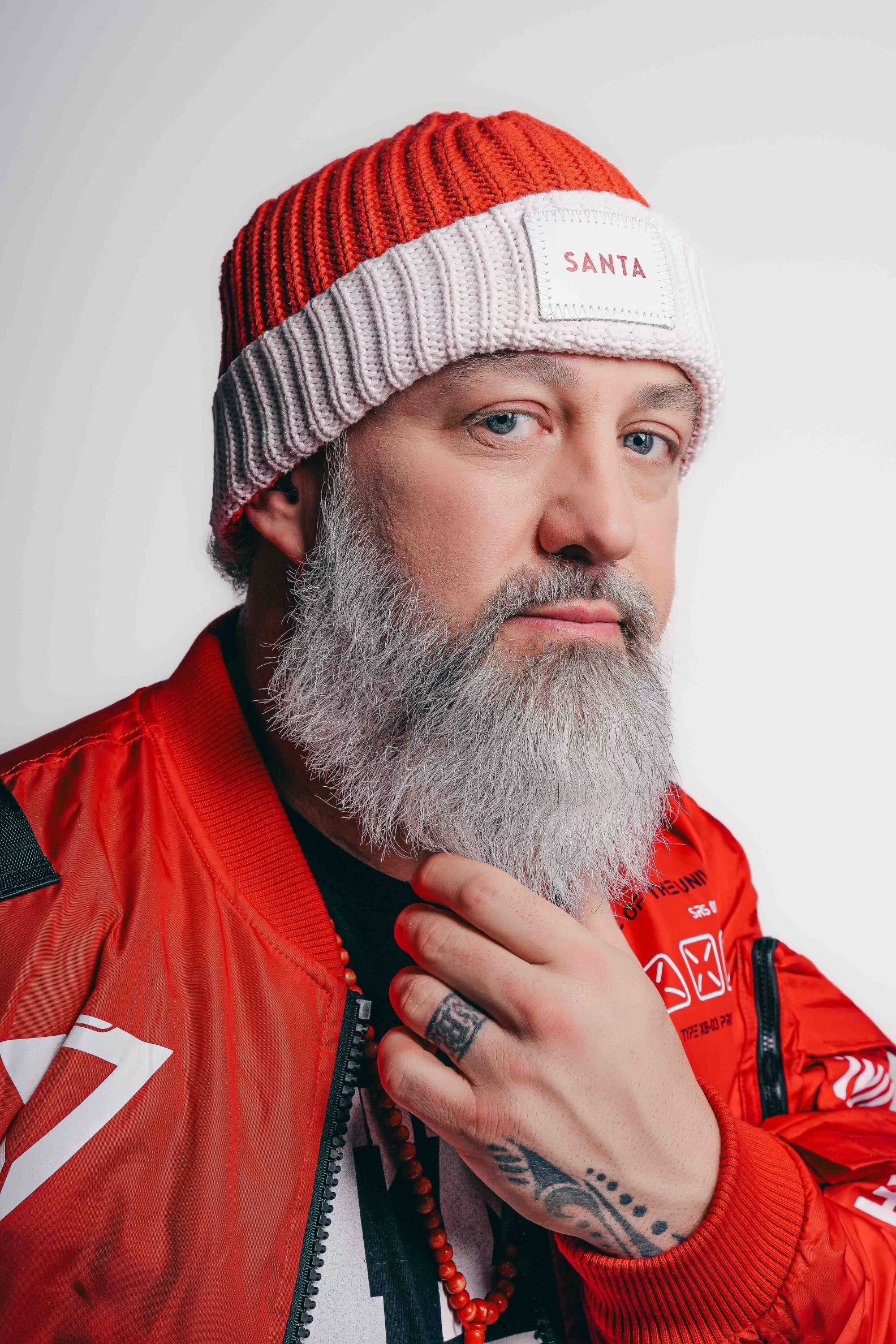 A man with a full white beard wearing a red and white beanie labeled 'Santa' on the front. He is dressed in a red jacket and is looking slightly to the side with a thoughtful expression, touching his beard with one hand. The background is plain white, highlighting the subject