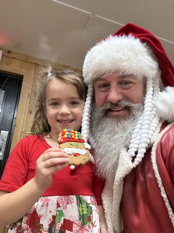 a middle aged man dressed as santa with a white beard and tassled hat standing next to a young girl in a christmas themed outfit holding a cookie in the shape of a santa head. they are both smiling standing in a white ceiling and brown wooden walls