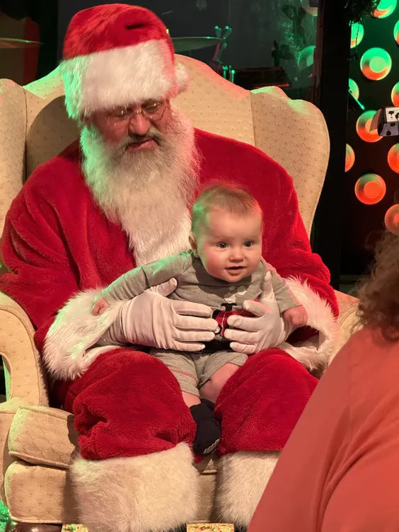 a middle aged man dressed as santa with a large white beard sitting on a cream colored plush chair. he has a baby on his lap smiling at his mother taking a picture. they are sitting in front of a drum kit and stage with dark walls