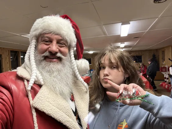 a middle aged man with a large beard and a red leather coat smiling next to a teenager in a blue hoodie. they are both standing in a room with a white roof and brown walls.