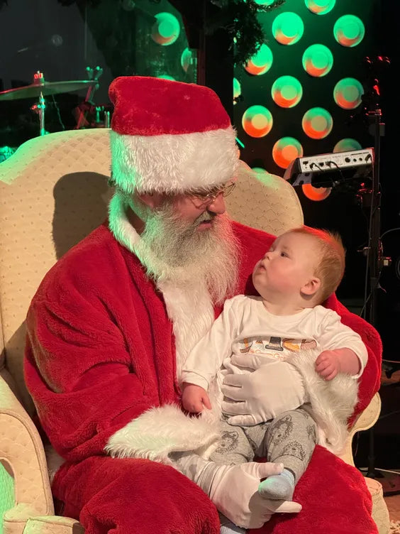 a middle aged man seated in a cream colored plush chair dressed as santa claus with a large white beard. there is a baby sitting on his lap and they are both seated in a room with dark walls, and a drum kit and guitar behind them.