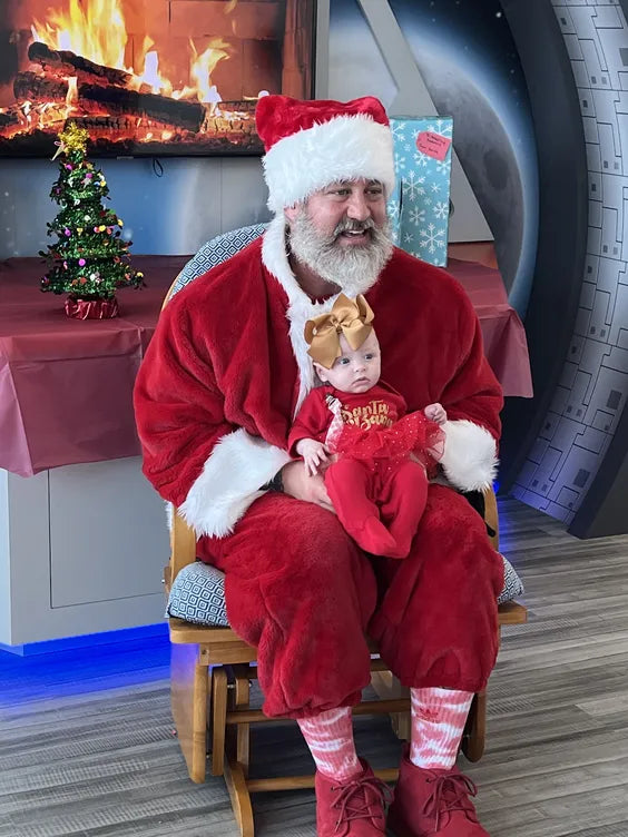 A middle aged man sitting in a wooden rocking chair in front of an artificial fireplace and space themed wall decoration. the man is dressed as santa claus and is smiling. he has a baby on his lap, also dressed in christmas clothing