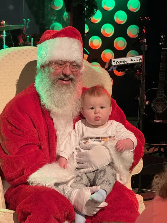 a middle aged man sitting in a cream colored plush chair dressed as santa claus with a large white beard. there is a baby sitting on his lap and they are both seated in a dark-walled room with a guitar and drum kit behind them