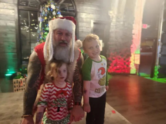 a middle aged man with a white beard and a tassled santa hat and christmas themed outfit kneeling behind two young children in christmas themed outfits standing in a christmas themed stone walled living room with red and green lights.