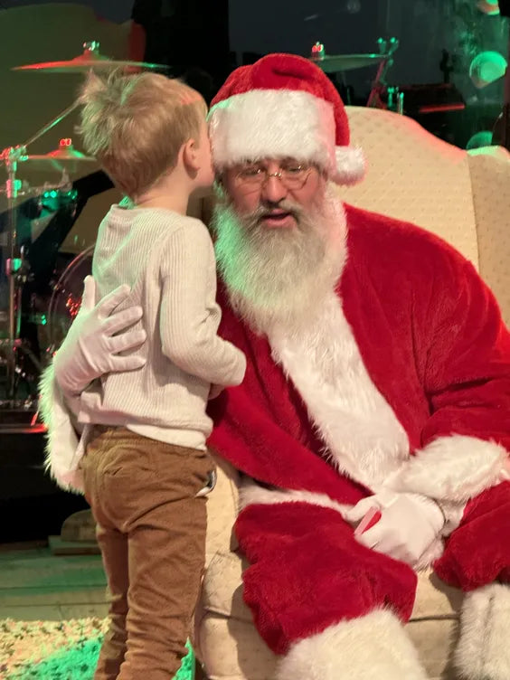 a middle aged man dressed as santa sitting in a cream colored plush chair leaning into a small boy whispering into his ear. they are sitting and standing in a room with a drum kit behind them and green lighting