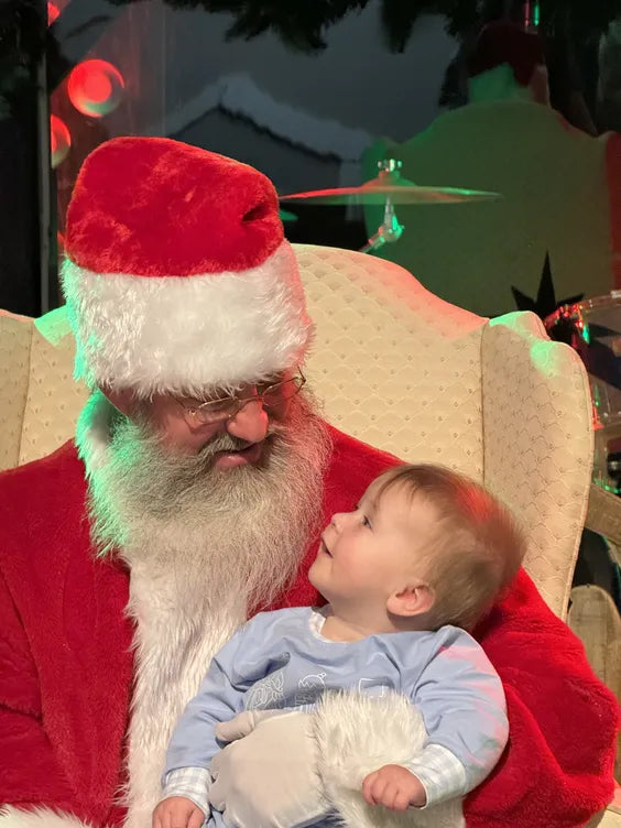 A middle aged man with a white beard dressed as santa claus sitting in a plush white chair looking down at a young toddler dressed in a baby blue onesie. they are sitting in an event room with a drum kit behind them, and a christmas themed backdrop