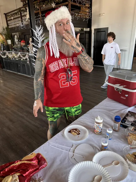 A middle aged man with a fully white beard and tattoos standing in an event room dressed in a christmas themed jersey licking his finger. the background is an event center with a loft, bar, and dark wood floors