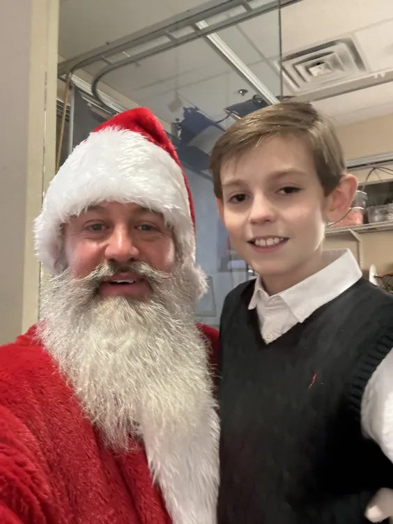 a middle aged man dressed as santa with his arm around a young boy in a collard white shirt and a black sweater vest. they are standing in a garage